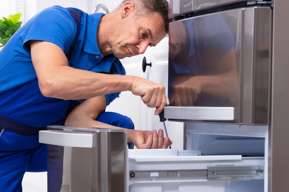 Mature,male,serviceman,repairing,refrigerator,with,toolbox,in,kitchen